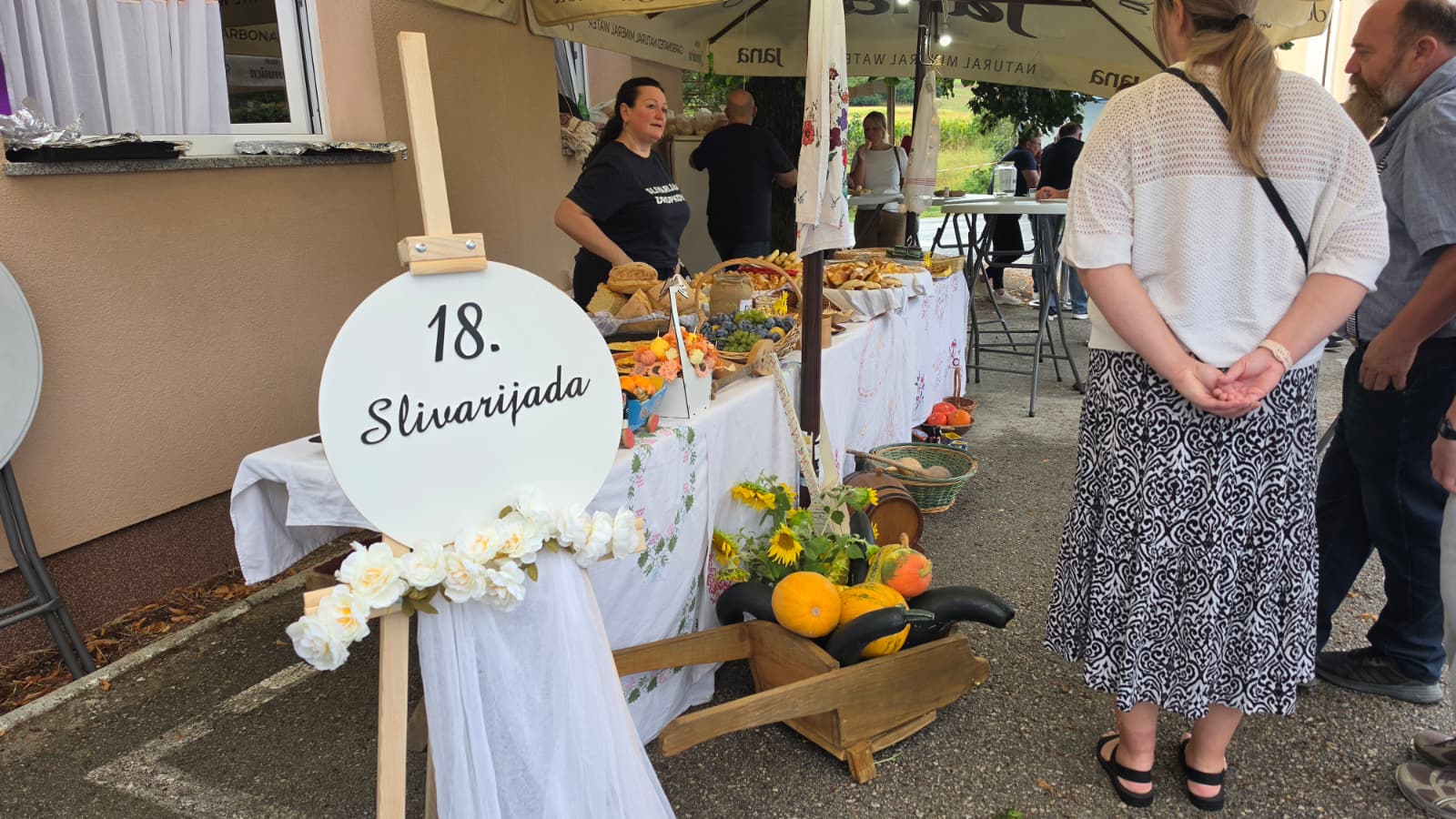 Ulaz na 18. Slivarijadu u Dropkovcu — bijela ploča s natpisom 18. Slivarijada ispred stola prepunog domaćih kruhova, kolača i jesenske dekoracije, uz goste pod šatorima ispred vatrogasnog doma
