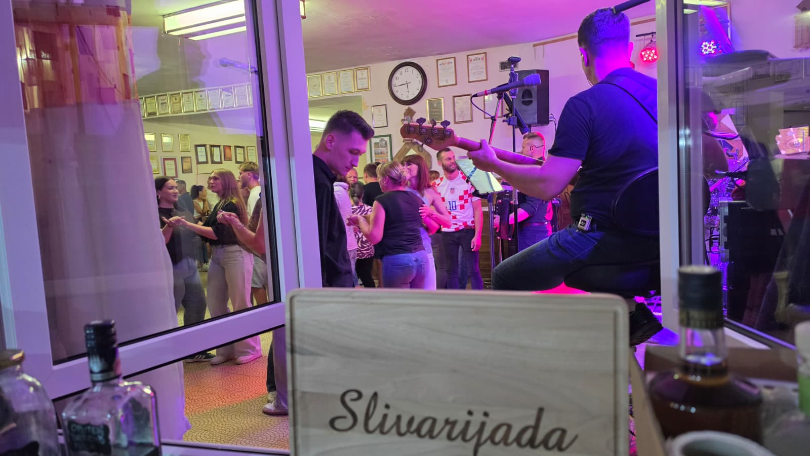 Pogled kroz prozor šanka na rasplesanu dvoranu i basista Joker Banda uz natpis Slivarijada
