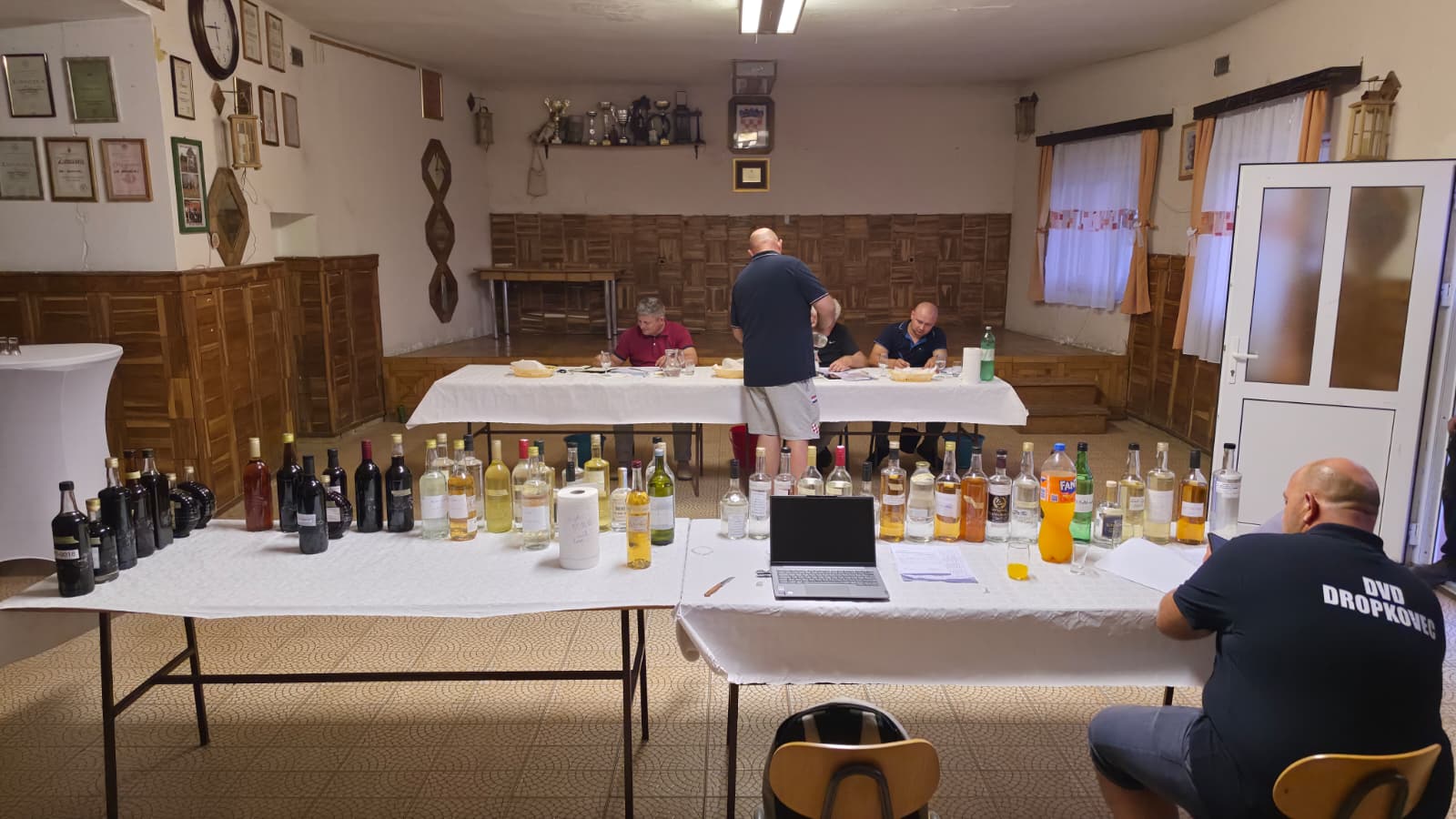 Ocjenjivanje rakija za 18. Slivarijadu u dvorani Vatrogasnog doma DVD-a Dropkovec — stolovi s numeriranim bocama uzoraka, stručno povjerenstvo za stolom u pozadini i član DVD-a Dropkovec s prijenosnim računalom