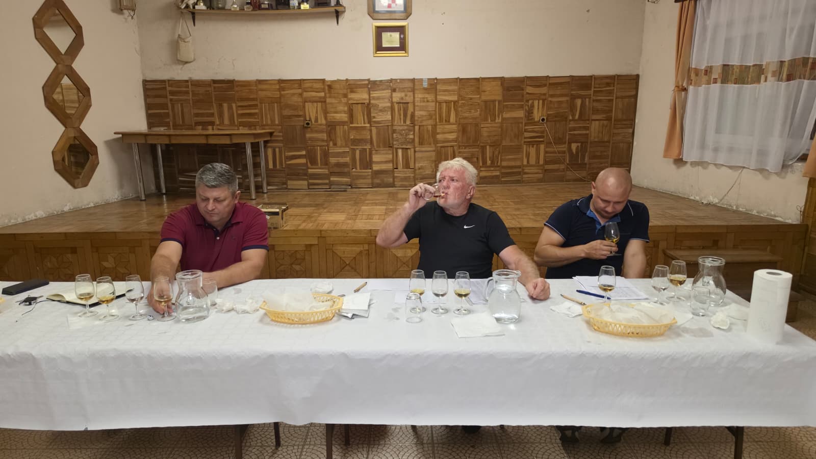 Tročlano stručno povjerenstvo za dugim stolom s bijelim stolnjakom kuša uzorke rakije na ocjenjivanju za 18. Slivarijadu